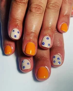 Short nails alternating solid orange and nude with tiny blue evil-eye dot designs.